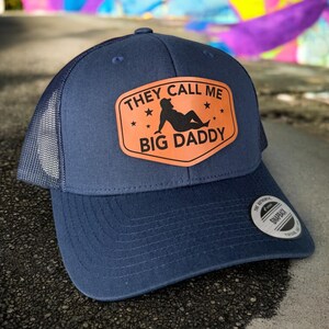 Op de afbeelding: Marineblauwe truckerpet met een mesh achterkant en een gebogen klep. De pet heeft een oranje patch met de tekst "THEY CALL ME BIG DADDY" en een silhouet. Een snapback sluiting is zichtbaar.