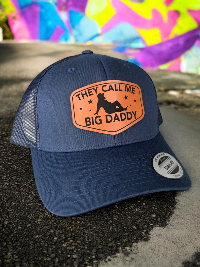 Op de afbeelding: Marineblauwe truckerpet met een mesh achterkant en een gebogen klep. De pet heeft een oranje patch met de tekst "THEY CALL ME BIG DADDY" en een silhouet. Een snapback sluiting is zichtbaar.