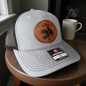 May include: A gray trucker hat with a black mesh back and a brown leather patch featuring a raven design and the text "F-CAW-F". The hat has a curved brim and an adjustable strap. The Richardson brand tag is visible.