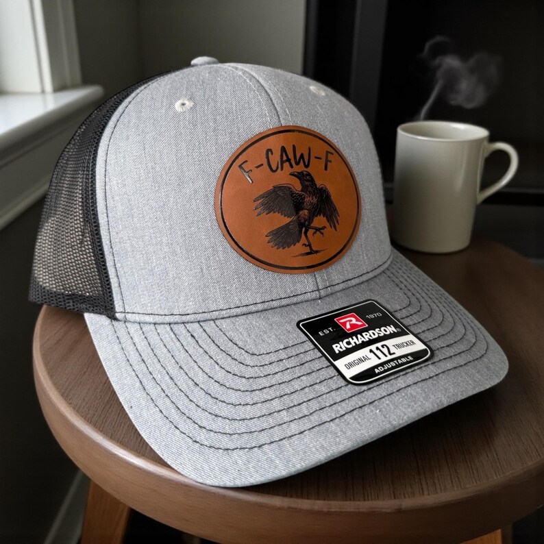 May include: A gray trucker hat with a black mesh back and a brown leather patch featuring a raven design and the text "F-CAW-F". The hat has a curved brim and an adjustable strap. The Richardson brand tag is visible.