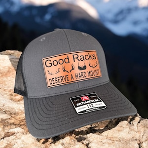 Pu&ograve; includere: Un cappello da camionista grigio con una rete nera sul retro. Il cappello ha una toppa in pelle marrone con il testo "Good Racks Deserve A Hard Mount" e una grafica di corna e un reggiseno. Il cappello ha anche un'etichetta nera e rossa con il testo "Richardson 112 Original Trucker Adjustable".