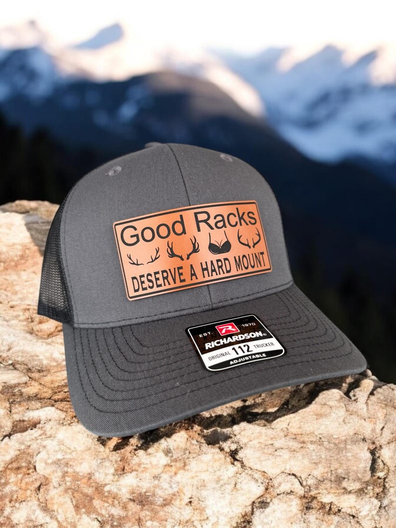 Pu&ograve; includere: Un cappello da camionista grigio con una rete nera sul retro. Il cappello ha una toppa in pelle marrone con il testo "Good Racks Deserve A Hard Mount" e una grafica di corna e un reggiseno. Il cappello ha anche un'etichetta nera e rossa con il testo "Richardson 112 Original Trucker Adjustable".