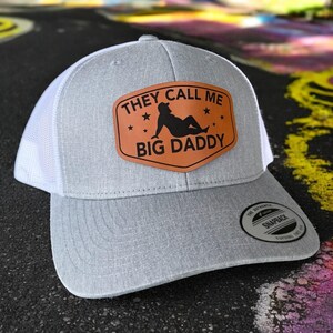 Op de afbeelding: Grijze en witte truckerpet met een gebogen klep. De voorkant heeft een bruin leren patch met de tekst "THEY CALL ME BIG DADDY" en een silhouet. Voorzien van een snapback sluiting.