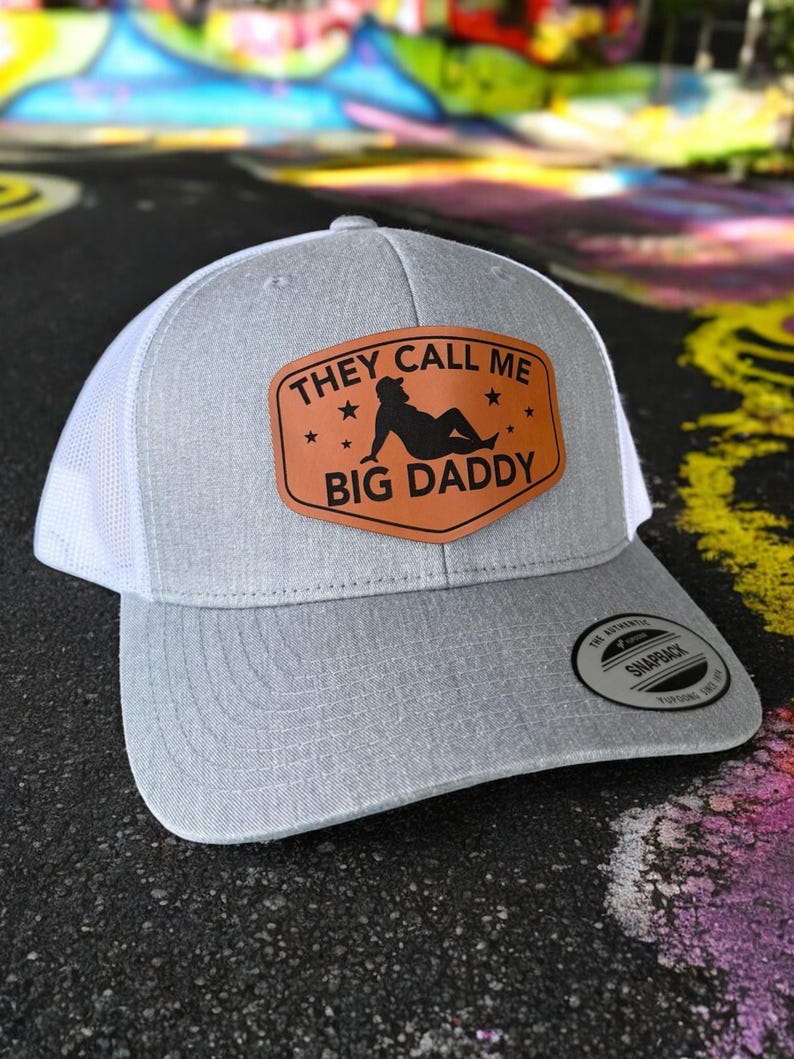 Op de afbeelding: Grijze en witte truckerpet met een gebogen klep. De voorkant heeft een bruin leren patch met de tekst "THEY CALL ME BIG DADDY" en een silhouet. Voorzien van een snapback sluiting.