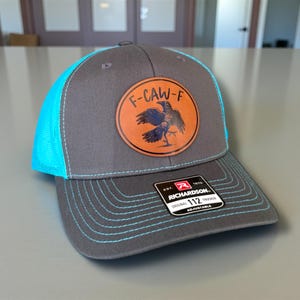 May include: Gray trucker hat with a teal mesh back and brim stitching. The hat features a circular leather patch with the text "F-CAW-F" and a raven illustration. The hat is adjustable and made by Richardson.