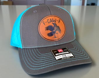 May include: Gray trucker hat with a teal mesh back and brim stitching. The hat features a circular leather patch with the text "F-CAW-F" and a raven illustration. The hat is adjustable and made by Richardson.