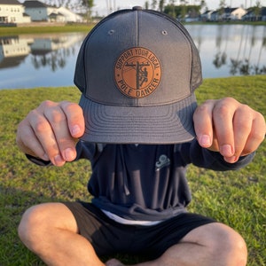 Può includere: Un cappello da camionista grigio e nero con una toppa in pelle marrone che dice "Support Your Local Pole Dancer". La toppa presenta una silhouette di una persona che scala un palo della luce.