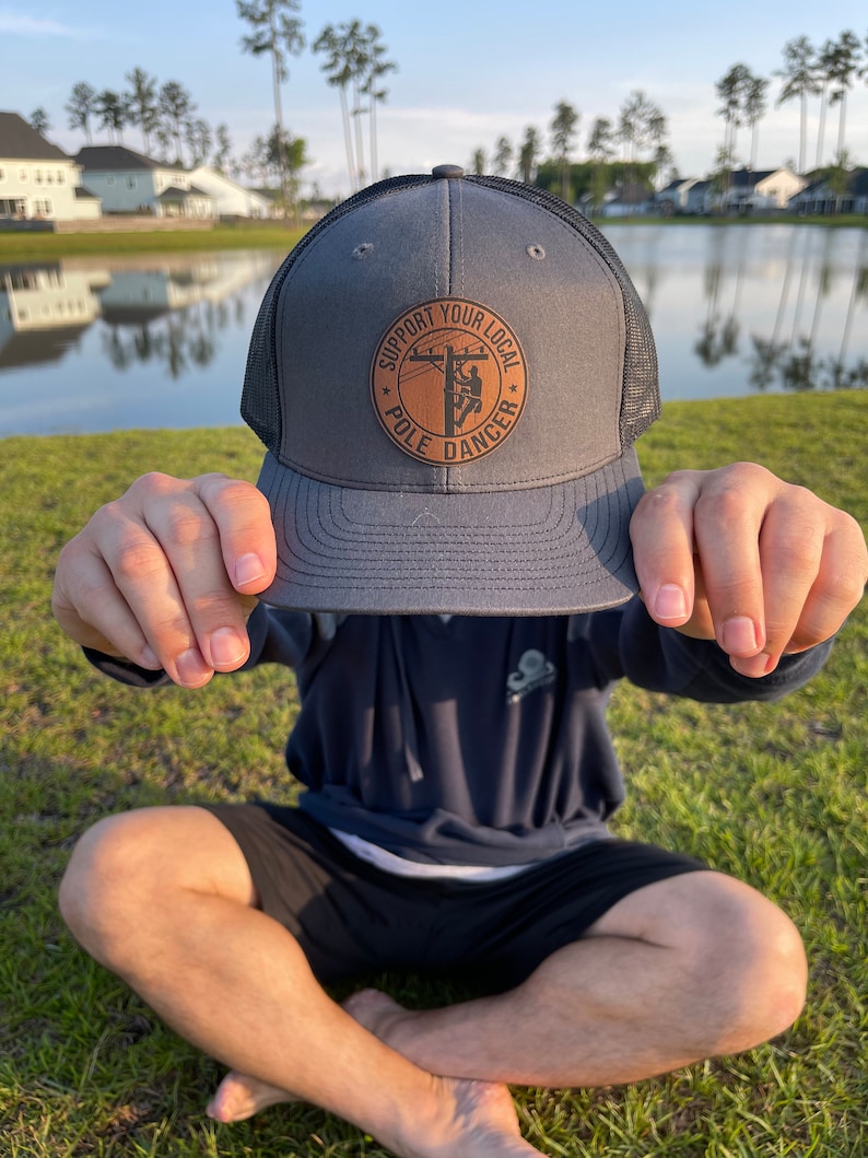 May include: A gray and black trucker hat with a brown leather patch that says "Support Your Local Pole Dancer".