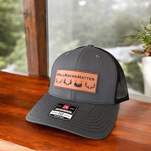 Pu&ograve; includere: Un cappello da camionista grigio e nero con una toppa in pelle marrone che dice "#ALLRACKSMATTER" con un palco di cervo e una grafica di reggiseno. Il cappello &egrave; regolabile e ha un logo Richardson.