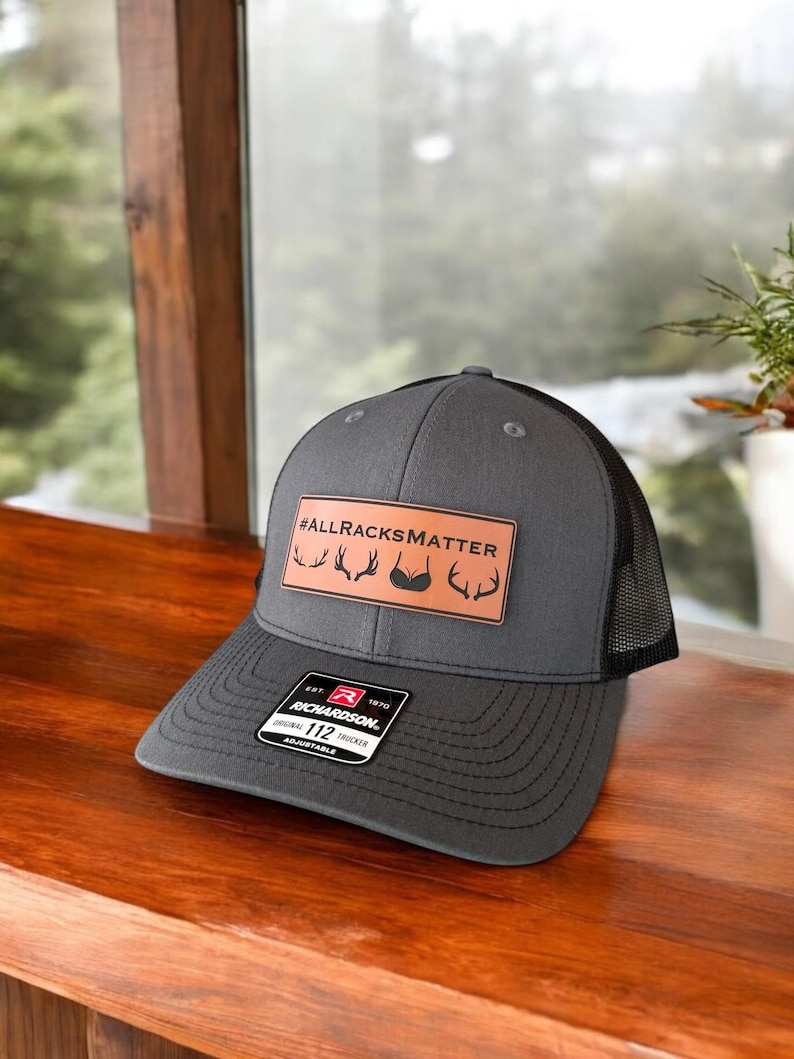 Pu&ograve; includere: Un cappello da camionista grigio e nero con una toppa in pelle marrone che dice "#ALLRACKSMATTER" con un palco di cervo e una grafica di reggiseno. Il cappello &egrave; regolabile e ha un logo Richardson.