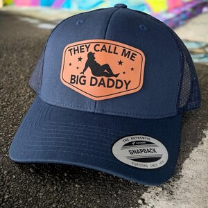 Op de afbeelding: Marineblauwe truckerpet met een gebogen klep en mesh achterkant. De pet heeft een rechthoekige patch met de tekst "THEY CALL ME BIG DADDY" en een silhouet van een persoon. Een "Yupoong"-label staat op de zijkant.