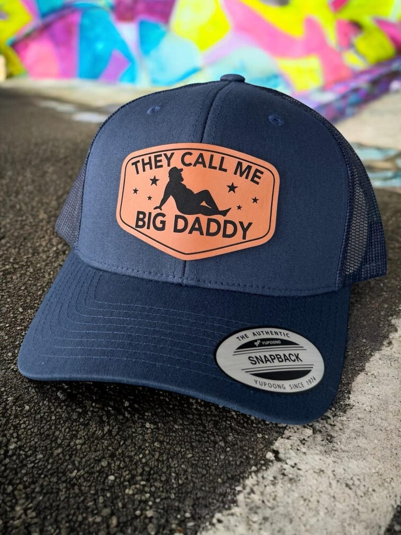 Op de afbeelding: Marineblauwe truckerpet met een gebogen klep en mesh achterkant. De pet heeft een rechthoekige patch met de tekst "THEY CALL ME BIG DADDY" en een silhouet van een persoon. Een "Yupoong"-label staat op de zijkant.