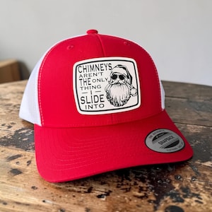 May include: A red and white trucker cap featuring a rectangular patch on the front. The patch displays a graphic of Santa Claus wearing sunglasses and the text: CHIMNEYS AREN'T THE ONLY THING I SLIDE INTO. A black circular snapback sticker is on the brim.