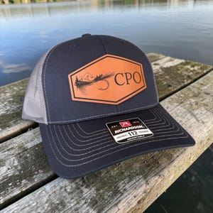 May include: A navy blue Richardson 112 trucker hat with a grey mesh back. The hat features a brown leather patch with a mountain and fishing hook design, and the letters "CPO". The hat is adjustable.