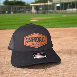 May include: Black and brown baseball cap with a mesh back. The front of the cap features a brown leather patch with the text "SOFTBALL COACH" in white lettering.