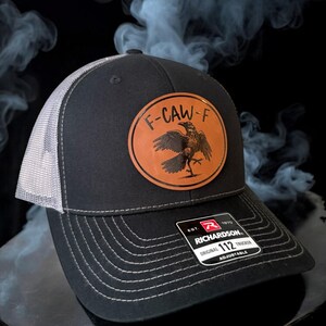 May include: Black trucker hat with a gray mesh back and a leather patch featuring a raven and the text "F-CAW-F". The hat is adjustable and has the Richardson 112 Trucker label.