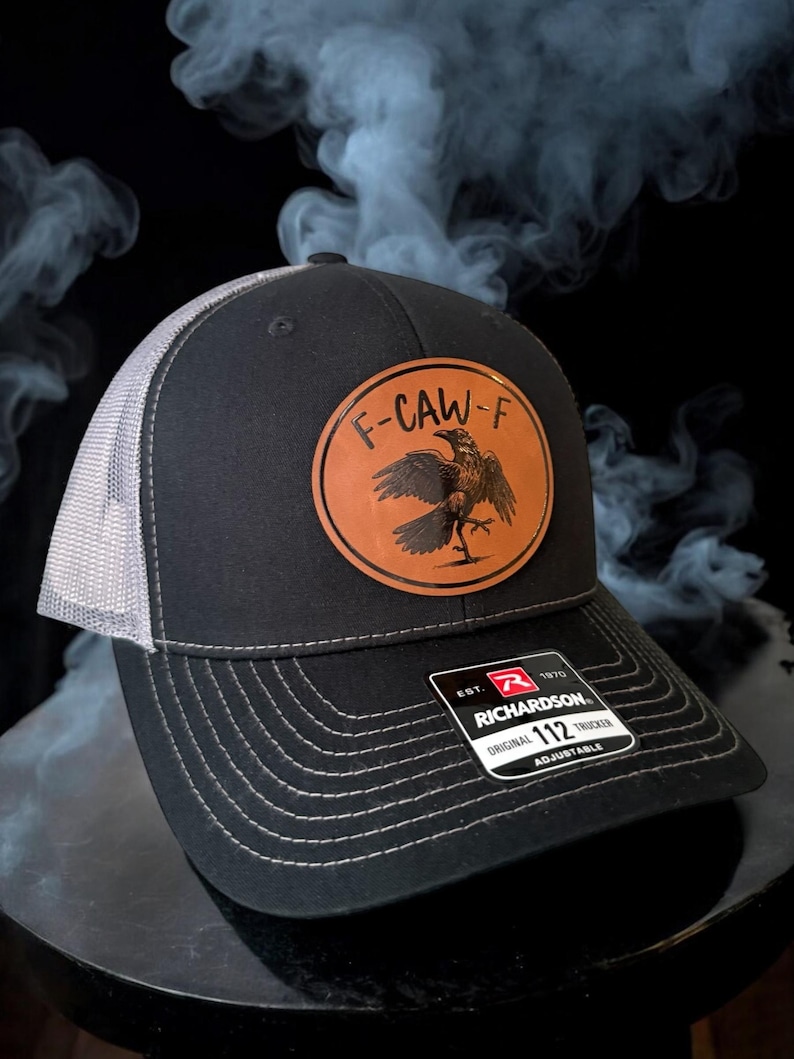 May include: Black trucker hat with a gray mesh back and a leather patch featuring a raven and the text "F-CAW-F". The hat is adjustable and has the Richardson 112 Trucker label.