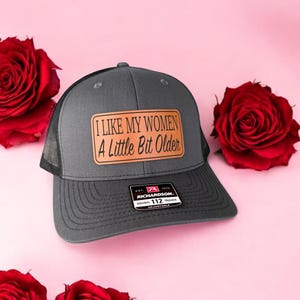 Puede incluir: Una gorra de camionero gris y negra con un parche de cuero marrón que dice "Me gustan mis mujeres un poco mayores".