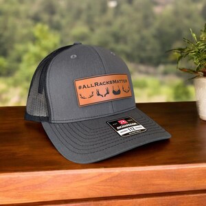 Pu&ograve; includere: Un cappello da camionista grigio e nero con una toppa in pelle marrone che dice "#ALLRACKSMATTER" con una grafica di corna di cervo. Il cappello &egrave; realizzato da Richardson 112.