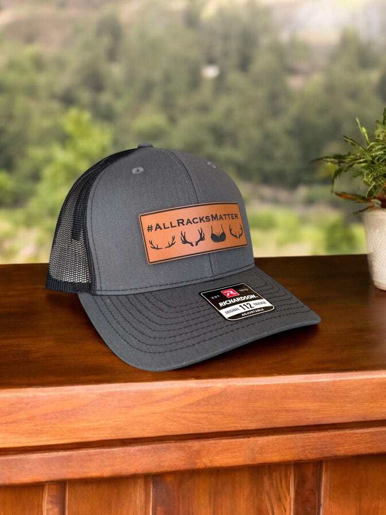 Pu&ograve; includere: Un cappello da camionista grigio e nero con una toppa in pelle marrone che dice "#ALLRACKSMATTER" con una grafica di corna di cervo. Il cappello &egrave; realizzato da Richardson 112.