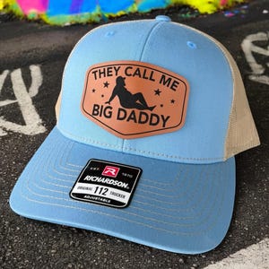 Op de afbeelding: Lichtblauwe Richardson 112 truckerpet met een beige mesh achterkant. De pet heeft een bruin leren patch met de tekst "THEY CALL ME BIG DADDY" en een silhouet van een persoon. Verstelbaar.