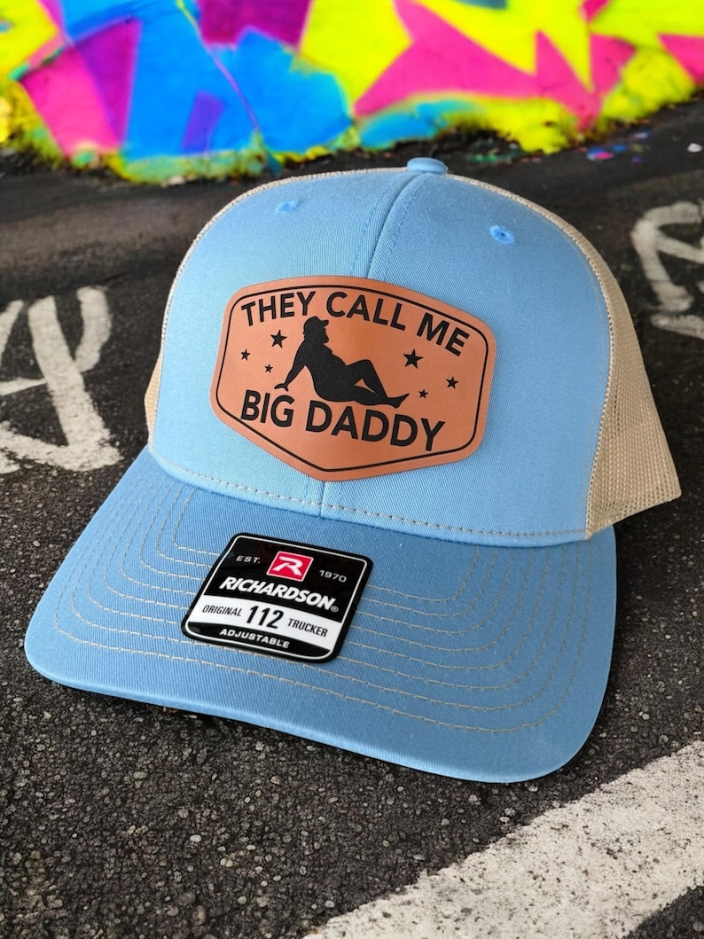 Op de afbeelding: Lichtblauwe Richardson 112 truckerpet met een beige mesh achterkant. De pet heeft een bruin leren patch met de tekst "THEY CALL ME BIG DADDY" en een silhouet van een persoon. Verstelbaar.