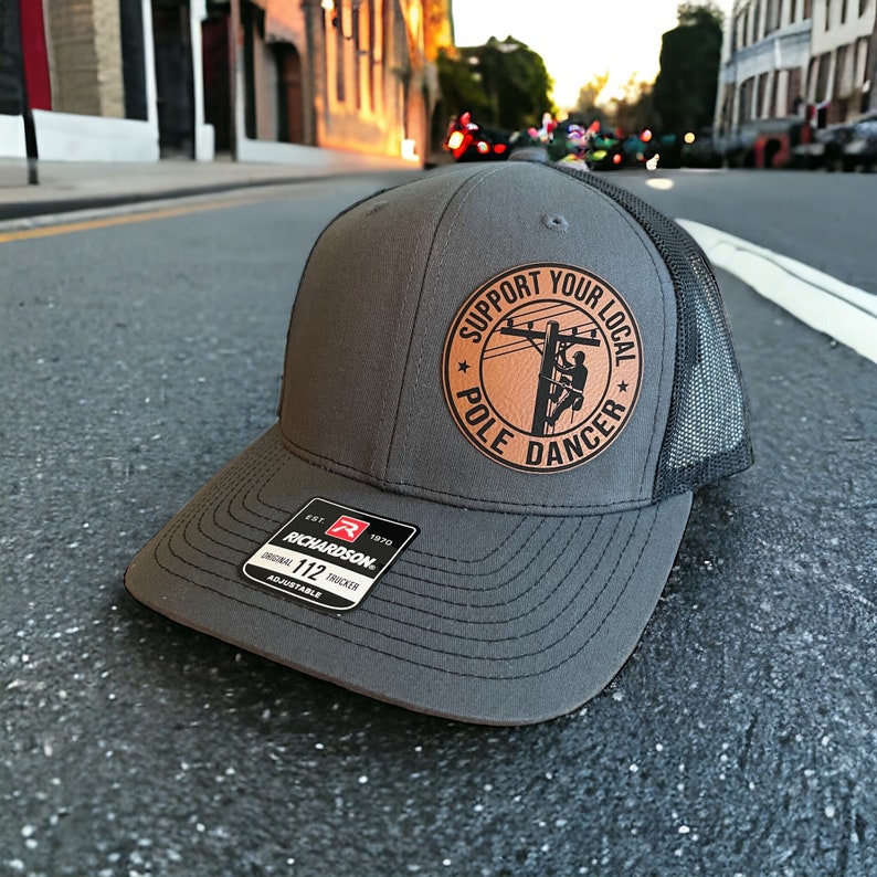 May include: A gray and black trucker hat with a brown leather patch that says "Support Your Local Pole Dancer". The patch features a silhouette of a person climbing a utility pole.