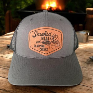 May include: Gray trucker hat with a mesh back and a curved brim. The hat features a brown leather patch with the words "Smokin' Meats and Clapping Cheeks" and a cartoon pig grilling.
