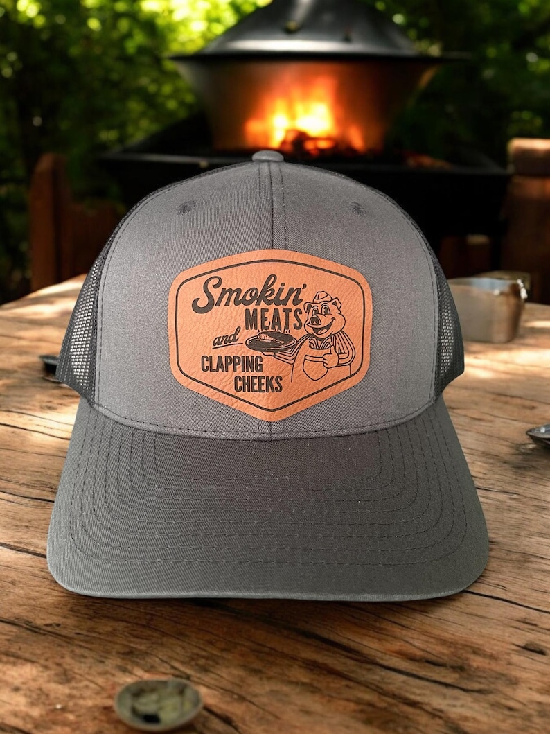 May include: Gray trucker hat with a mesh back and a curved brim. The hat features a brown leather patch with the words "Smokin' Meats and Clapping Cheeks" and a cartoon pig grilling.