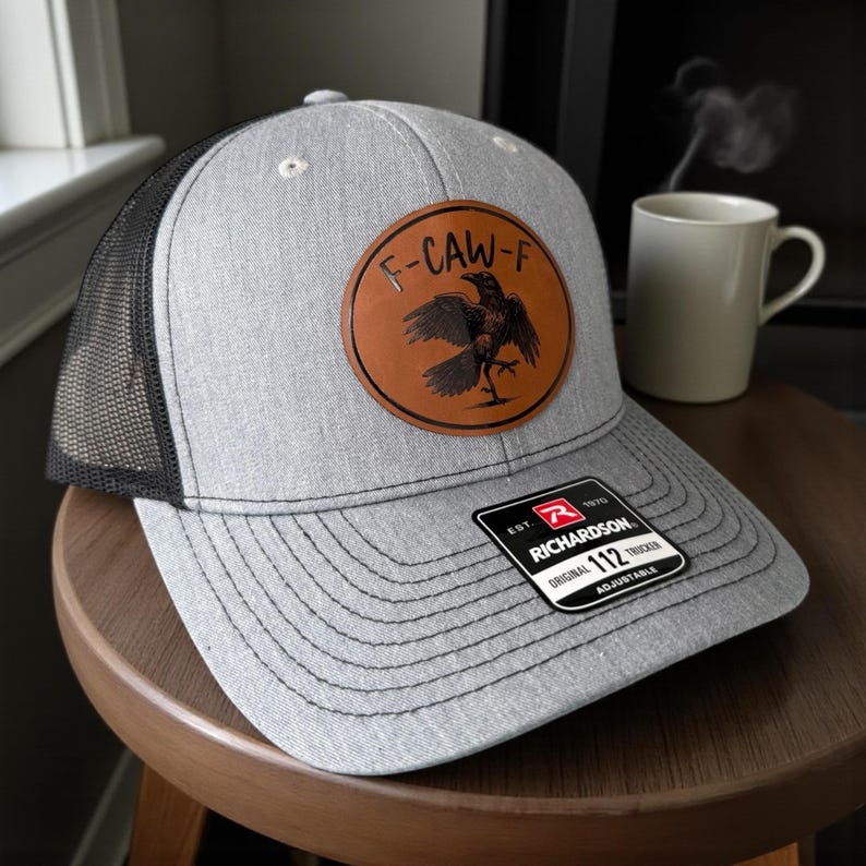 May include: A gray trucker hat with a black mesh back and a brown leather patch featuring a raven design and the text "F-CAW-F". The hat has a curved brim and an adjustable strap. The Richardson brand tag is visible.