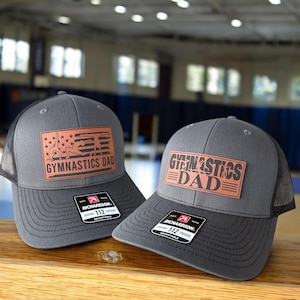 May include: Two gray trucker hats with black mesh backs and curved visors. Each hat has a rectangular patch with the words "GYMNASTICS DAD" and a gymnastics-themed design in orange. The hats are adjustable.