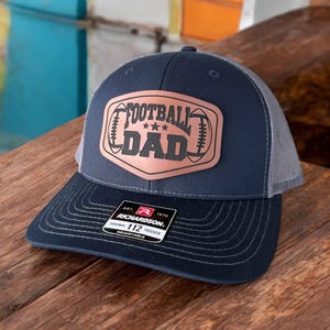 May include: Navy and grey trucker cap featuring a copper-toned patch with the words "FOOTBALL DAD". The cap has a curved peak and an adjustable snap closure. The Richardson brand label is visible.