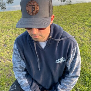 May include: A gray and black trucker hat with a brown leather patch that says "Support Your Local Pole Dancer". The patch features a silhouette of a person climbing a utility pole.