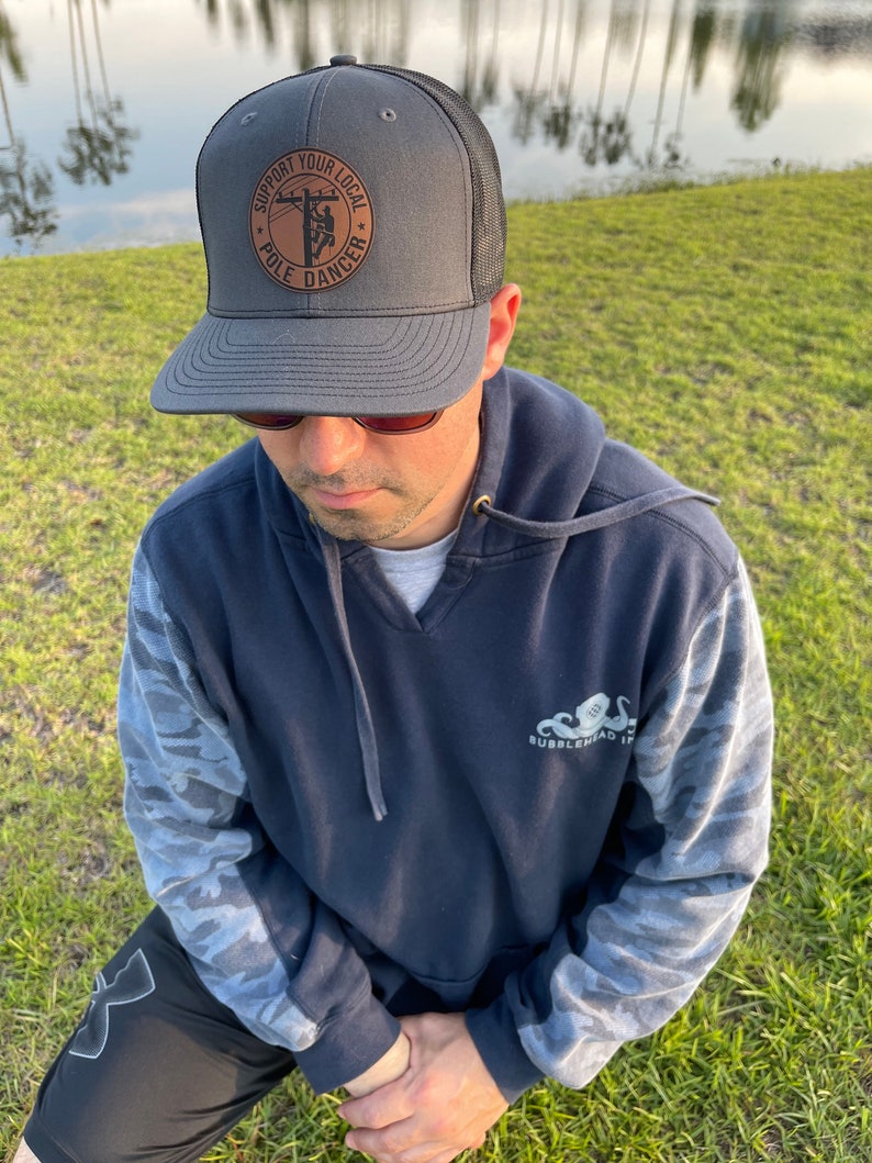 May include: A gray and black trucker hat with a brown leather patch that says "Support Your Local Pole Dancer". The patch features a silhouette of a person climbing a utility pole.