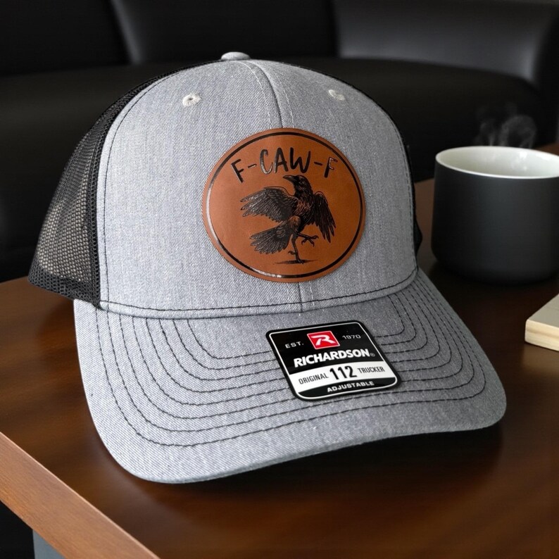May include: Gray trucker hat with a black mesh back and a brown leather patch featuring a crow graphic and the text "F-CAW-F". The hat has a curved brim and an adjustable strap. The tag reads "RICHARDSON 112 TRUCKER".