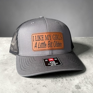 Puede incluir: Gorra de camionero gris con parte trasera de malla y visera curva. La gorra presenta un parche rectangular con el texto "I LIKE MY GIRLS A Little Bit Older". La etiqueta dice "112 The Original MD-LG".