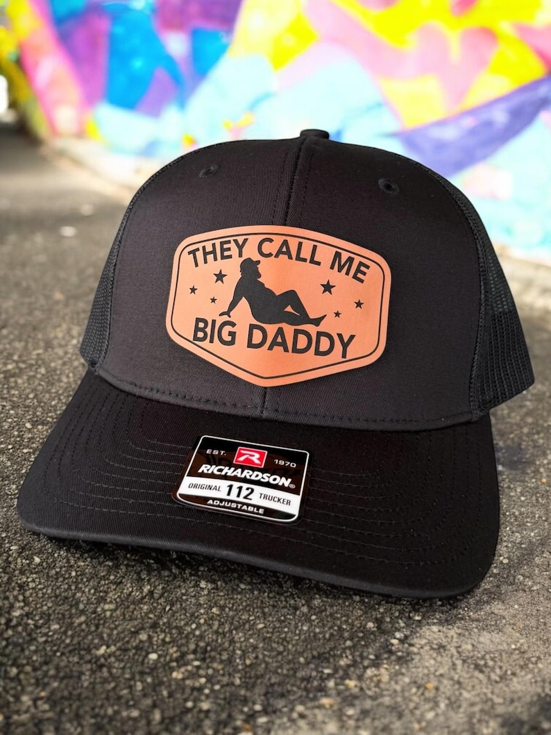 Op de afbeelding: Zwarte truckerpet met een mesh achterkant en een gebogen klep. De pet heeft een bruine patch met de tekst "THEY CALL ME BIG DADDY" en een silhouet. De pet is verstelbaar en heeft een Richardson 112 Trucker label.
