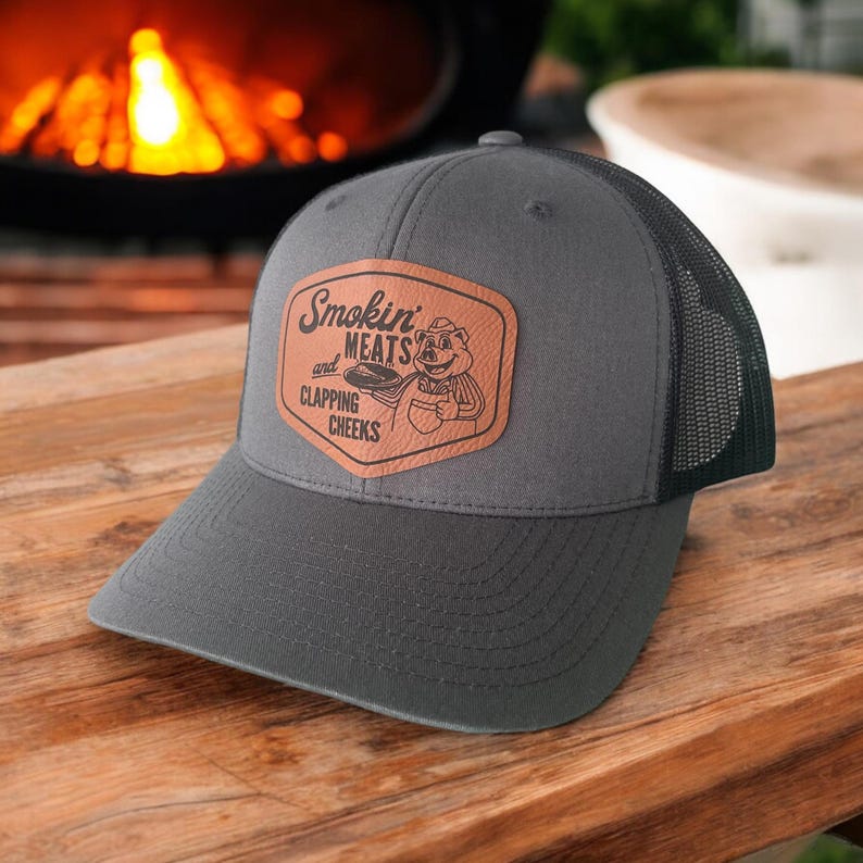 May include: Gray trucker hat with a black mesh back and a brown leather patch. The patch features the text "Smokin' MEATS and CLAPPING CHEEKS" with a cartoon pig graphic. The hat is on a wooden surface.