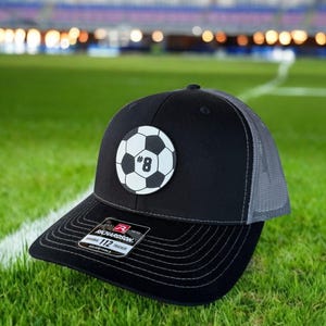 May include: Black and gray trucker hat with a soccer ball patch featuring the number 8. The hat has a curved brim and a mesh back. The Richardson brand label is visible on the brim.