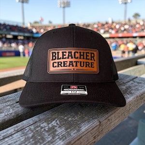May include: Black trucker hat with a mesh back and a rectangular patch that reads "BLEACHER CREATURE" in white letters on a brown background. The hat has a curved brim and an adjustable strap. The hat is sitting on a wooden surface.