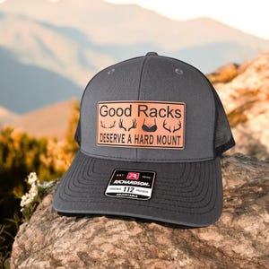 Pu&ograve; includere: Un cappello da camionista grigio scuro con una toppa in pelle marrone che recita "Good Racks Deserve A Hard Mount". La toppa presenta un'immagine stilizzata di corna di cervo e un reggiseno. Il cappello &egrave; regolabile e ha un logo Richardson.