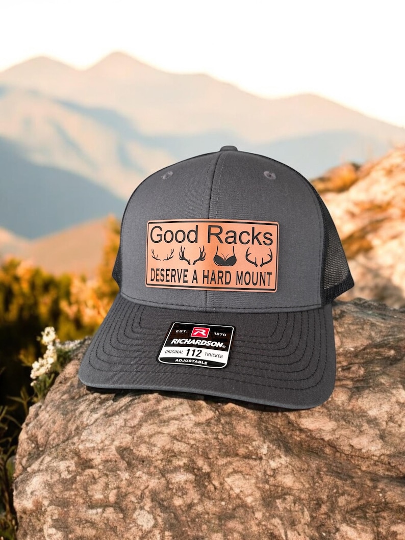 Pu&ograve; includere: Un cappello da camionista grigio scuro con una toppa in pelle marrone che recita "Good Racks Deserve A Hard Mount". La toppa presenta un'immagine stilizzata di corna di cervo e un reggiseno. Il cappello &egrave; regolabile e ha un logo Richardson.