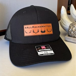 May include: Black trucker hat with a mesh back and a curved peak. The hat features a rectangular patch with the text "#ALLRACKSMATTER" and deer antlers and a bra graphic. It has an adjustable strap and a Richardson tag.