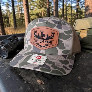 May include: A camouflage baseball cap featuring a tan leather-like patch on the front. The patch displays a black silhouette of a deer with antlers, pine trees, and two rifles, with the text CUSTOM NAME. A white sticker on the brim reads 112PFP.