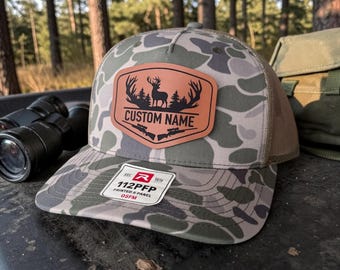 May include: A camouflage baseball cap featuring a tan leather-like patch on the front. The patch displays a black silhouette of a deer with antlers, pine trees, and two rifles, with the text CUSTOM NAME. A white sticker on the brim reads 112PFP.