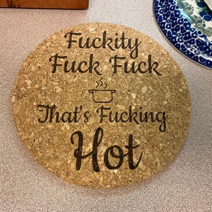 Puede incluir: Un posavasos redondo de corcho con el texto "F*ckity F*ck F*ck That's F*cking Hot" en letras negras. El posavasos tiene una pequeña ilustración de una olla con vapor saliendo de ella.