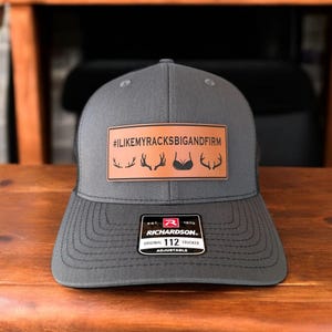 Pu&ograve; includere: Un cappello da camionista grigio con una toppa in pelle marrone che dice "#ILIKEMYRACKSBIGANDFIRM" con una grafica di corna di cervo. Il cappello &egrave; regolabile e ha un logo Richardson 112.
