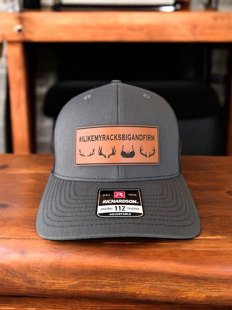 Pu&ograve; includere: Un cappello da camionista grigio con una toppa in pelle marrone che dice "#ILIKEMYRACKSBIGANDFIRM" con una grafica di corna di cervo. Il cappello &egrave; regolabile e ha un logo Richardson 112.