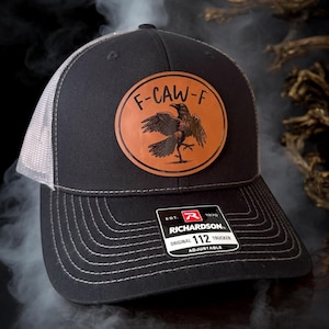 May include: Black trucker hat with a gray mesh back and a curved brim. The hat features a circular brown leather patch with a raven illustration and the text "F-CAW-F". A tag on the brim reads "RICHARDSON 112 TRUCKER".