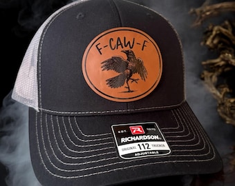 May include: Black trucker hat with a gray mesh back and a curved brim. The hat features a circular brown leather patch with a raven illustration and the text "F-CAW-F". A tag on the brim reads "RICHARDSON 112 TRUCKER".